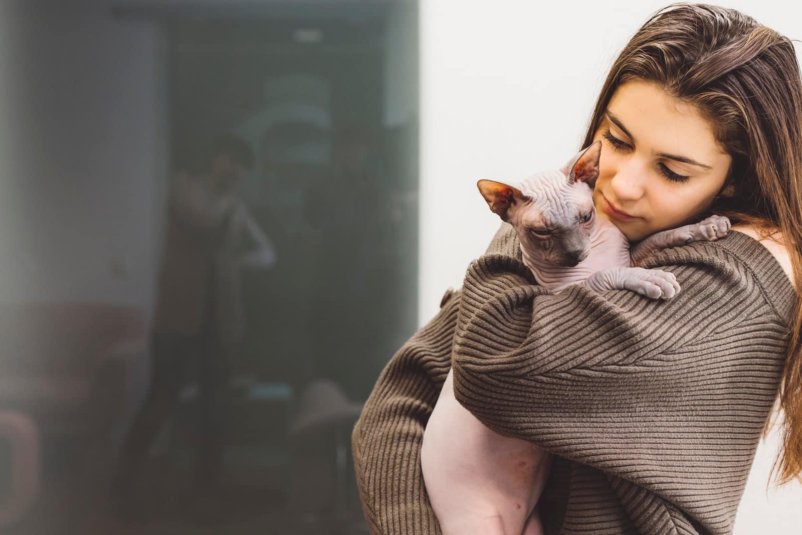 A young woman gently holds a Sphynx cat in her arms, showcasing a tender moment. The woman has long